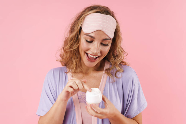 Изображение белой веселой женщины в маске для сна, улыбающейся при нанесении крема для лица на розовый фон
 - Фото, изображение