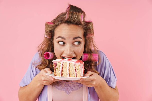 Image of young happy woman in curlers looking aside and eating cake isolated over pink background - Foto, Bild