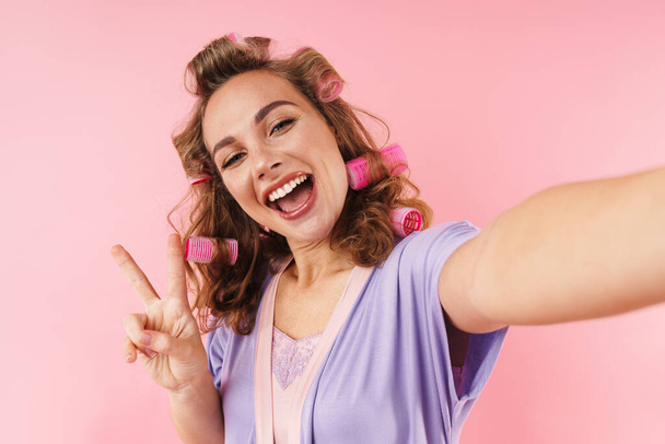 Imagem da jovem mulher sorridente em curlers gesticulando sinal de paz ao tomar selfie isolado sobre fundo rosa
 - Foto, Imagem