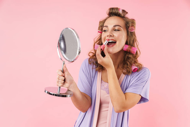 Bild einer zufriedenen jungen Frau in Lockenwicklern, die Lippenstift aufträgt und den Spiegel isoliert auf rosa Hintergrund betrachtet - Foto, Bild