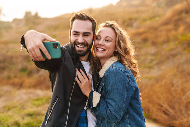 Beautiful young happy stylish couple wearing jackets standing at the beach, taking a selfie - 写真・画像