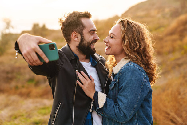 Beautiful young happy stylish couple wearing jackets standing at the beach, taking a selfie - Foto, Bild