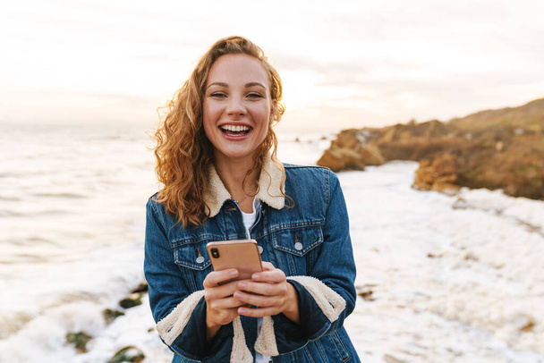 Image of young beautiful woman with blond curly hair using smartphone while walking by seaside - Photo, image