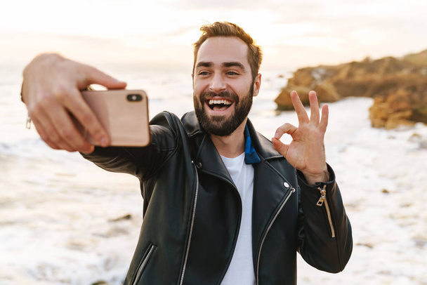 Image of young handsome man in leather jacket taking selfie photo on smartphone while walking by seaside - Photo, image