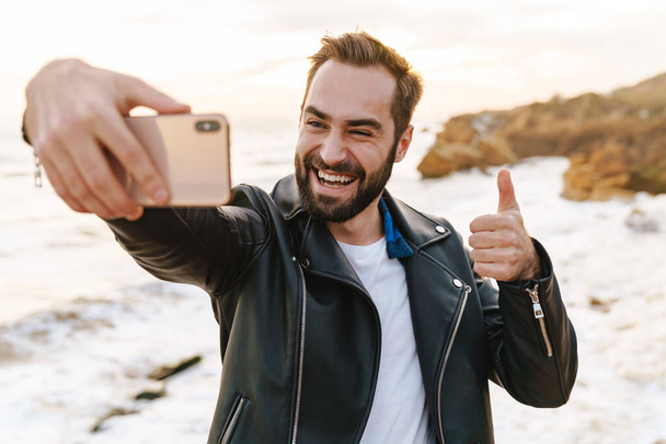 Image of young handsome man in leather jacket taking selfie photo on smartphone while walking by seaside - Фото, изображение