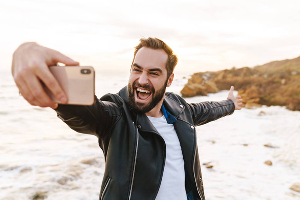 Image of young handsome man in leather jacket taking selfie photo on smartphone while walking by seaside - Zdjęcie, obraz