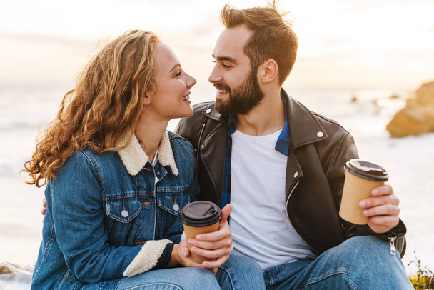 Image of beautiful young caucasian couple drinking coffee together while walking by seaside - Photo, image