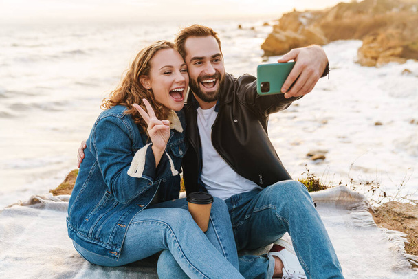 Image of beautiful young caucasian couple taking selfie photo on cellphone while walking by seaside - Foto, imagen