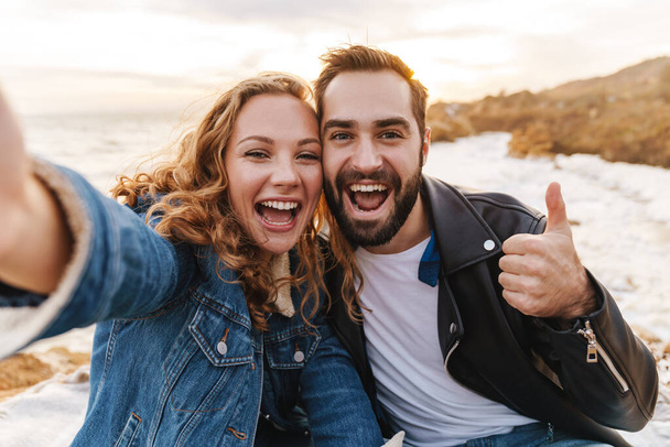 Image of beautiful young caucasian couple smiling and taking selfie photo while walking by seaside - Foto, imagen