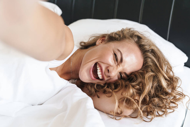 Close up of a happy beautiful young blonde haired woman laying on a pillow in bed, taking a selfie - Photo, Image