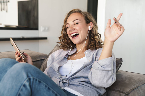 Cheerful young blonde haired woman listening to music with headphones while sitting on a couch at home and holding mobile phone - Foto, afbeelding