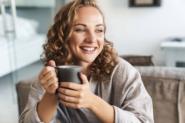 Cheerful young blonde haired woman drinking tea while sitting on a couch at home, looking away - Photo, image
