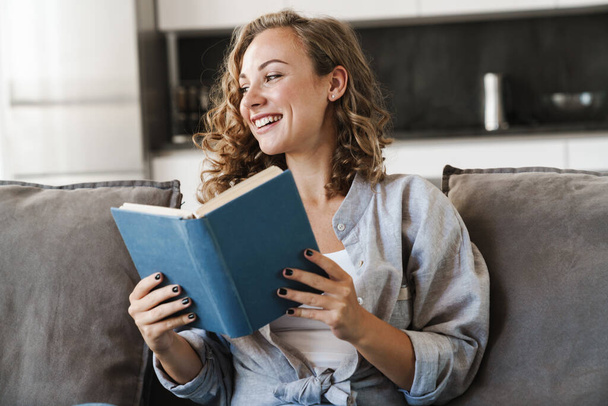 Cheerful young blonde haired woman reading a book while sitting on a couch at home - 写真・画像