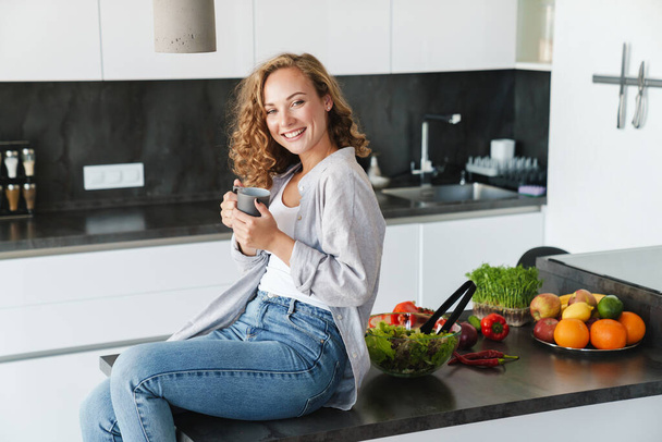 Smiling young woman wearing casual clothes having cup of coffee while sitting on a kitchen table - Foto, imagen