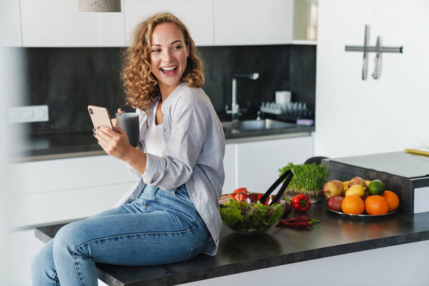 Smiling young woman wearing casual clothes having cup of coffee while sitting on a kitchen table and using mobile phone - Foto, Imagem