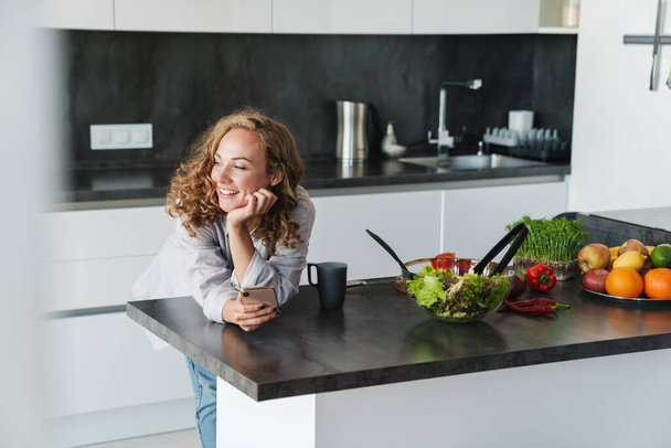 Smiling young woman wearing casual clothes using mobile phone while leaning on a kitchen table - Foto, Imagem