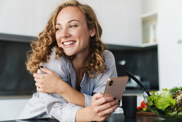 Mujer joven sonriente usando ropa casual usando teléfono móvil mientras se apoya en una mesa de cocina
 - Foto, Imagen