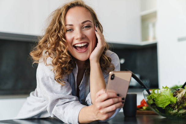 Mujer joven sonriente usando ropa casual usando teléfono móvil mientras se apoya en una mesa de cocina
 - Foto, Imagen
