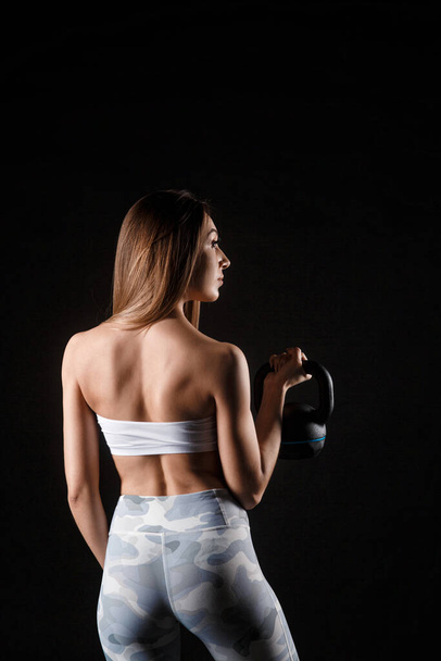 Fitness exercise woman holding kettlebell strength training biceps. Beautiful sweaty fitness instructor looking. Mixed race Caucasian female sweating isolated on black background. - Фото, зображення