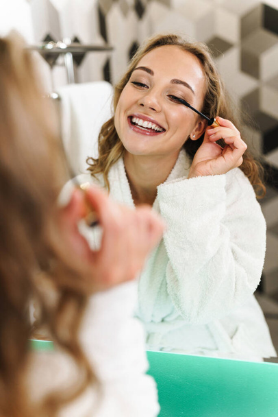 Imagen de la joven hermosa mujer rubia en la casa aplicando maquillaje después de la ducha por espejo en el baño
 - Foto, imagen
