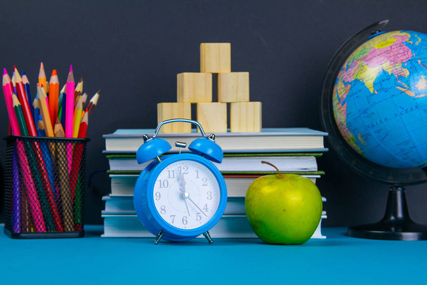 Una pila de libros con un globo, un vaso de lápices, un reloj y manzanas
. - Foto, Imagen