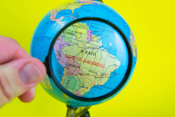 Close-up of a hand holding a magnifying glass to look at the globe in the library selective focus Brazil. - Photo, Image