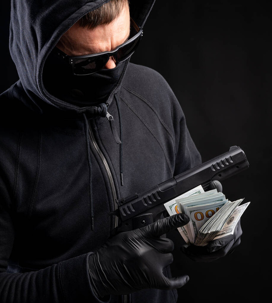 Άντρας με μαύρη μάσκα και κουκούλα με όπλο και πακέτο δολάρια πάνω από μαύρο φόντο. - Φωτογραφία, εικόνα