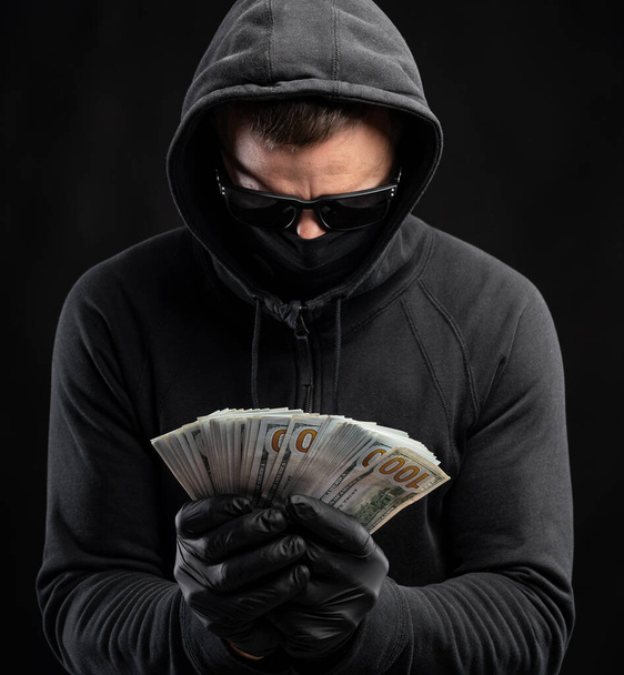 Η έννοια των οικονομικών. Άντρας με μαύρη μάσκα και κουκούλα να κρατάει ένα πακέτο δολάρια πάνω από μαύρο φόντο - Φωτογραφία, εικόνα