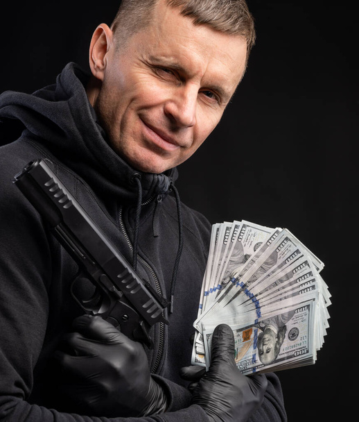 Uomo con cappuccio nero con pistola e pacchetto di dollari su sfondo nero
 - Foto, immagini