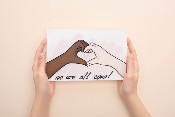 cropped view of woman holding picture with drawn multiethnic hands showing heart gesture on beige background - Photo, Image