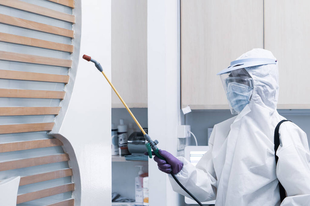 werknemer in persoonlijke beschermingsmiddelen, waaronder masker in wit pak en ontsmettingsmiddel ter bestrijding van coronavirusinfectie in de tandartsenkliniek - Foto, afbeelding