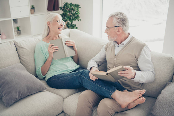 Portrait of cheerful grandmother grandfather hold fiction book laugh wear eyeglasses eyewear denim jeans trousers teal brown pullover sit lie divan bare foot in room - 写真・画像