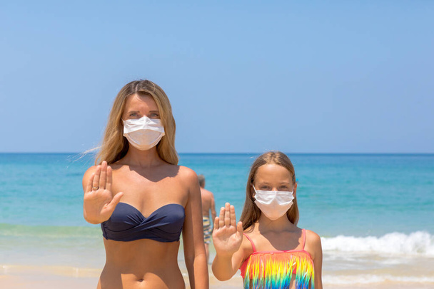 A woman and a child in a protective surgical mask on their face are standing on the beach in swimsuits. Chinese coronavirus disease COVID-19 is dangerous. - Foto, Imagen