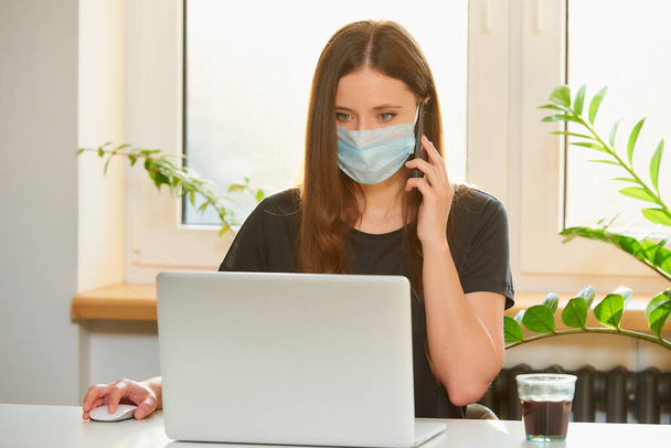 A woman in a medical face mask working remotely on her laptop during the quarantine to avoid the spread coronavirus. A lady calling to her business partner from home during the pandemic of COVID-19. - Foto, Imagem