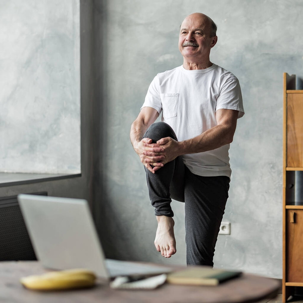 Senior deportista guapo haciendo estiramiento y equilibrio de ejercicio de yoga en casa utilizando la lección en línea en el portátil. Cómo mantenerse saludable con el concepto de cuarentena
. - Foto, Imagen