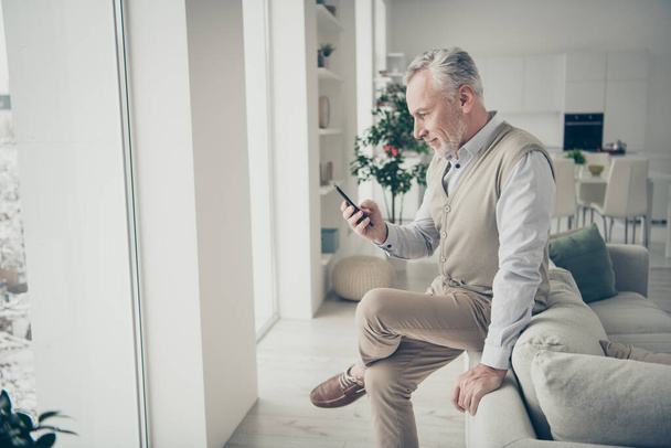 Photo of white haired aged man holding hand telephone searching news sitting sofa indoors - Photo, image