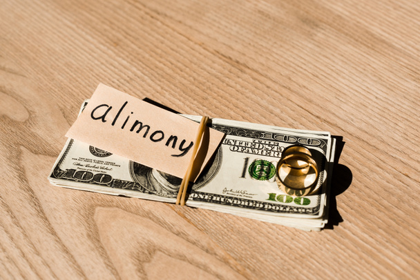 paper with alimony lettering on dollar banknotes near engagement rings  - Фото, изображение