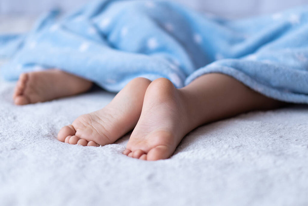 Heels and toes on bed. Heels and feets. Childrens feet. Barefeet on the bed. Kids feet in bed. Kids taking a rest focus on barefeet. Childrens bare feet look out from under the blue blanket. Bare - Photo, Image