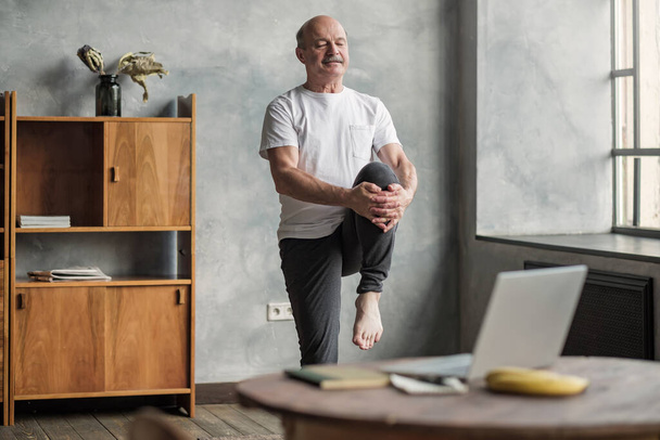 Senior knappe sportman doet stretching en balancing yoga oefening thuis met behulp van online les op notebook. Hoe gezond te blijven op quarantaine concept. - Foto, afbeelding