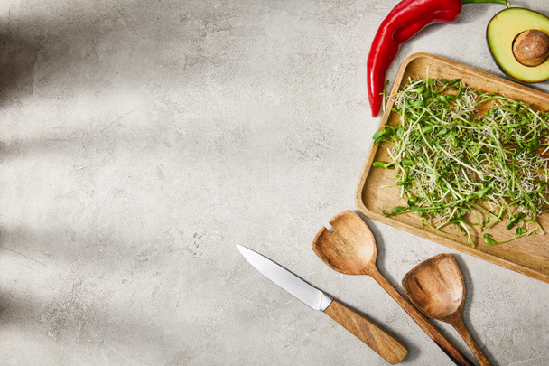 Top kilátás vágódeszka mikrozöldek, avokádó fele, chili paprika, spatulák és kés szürke háttér - Fotó, kép