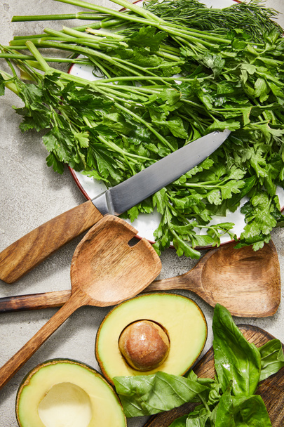 Top view of greenery on plate, knife, spatulas, avocado halves and basil leaves on grey background - 写真・画像