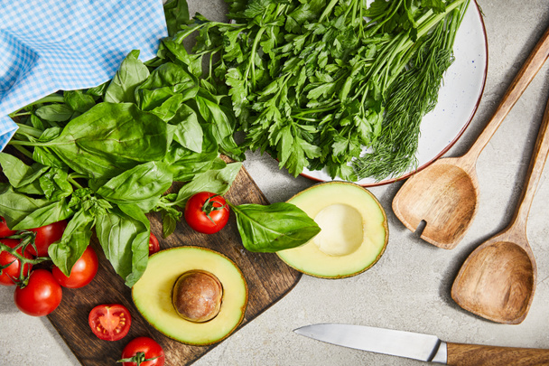 Top view of greenery, basil leaves, cherry tomatoes and avocado halves near spatulas, knife and cloth on grey background - Photo, Image