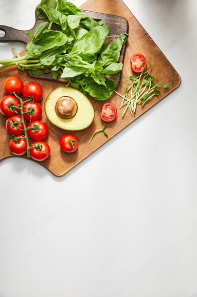 Top view of basil leaves, cherry tomatoes, avocado half and microgreens on cutting boards on white background - Photo, Image