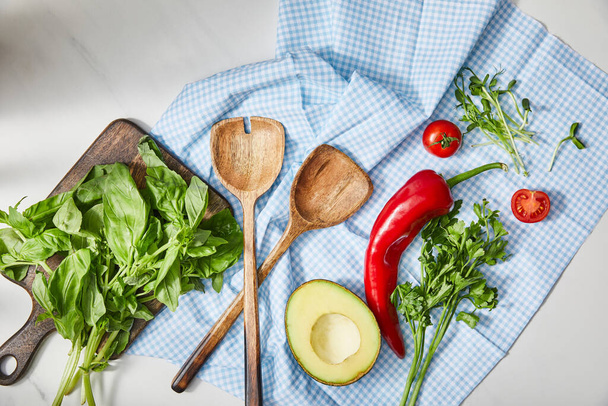 Top view of spatulas, parsley, microgreens, vegetables and avocado half on plaid cloth  near cutting board with basil on white  - Zdjęcie, obraz
