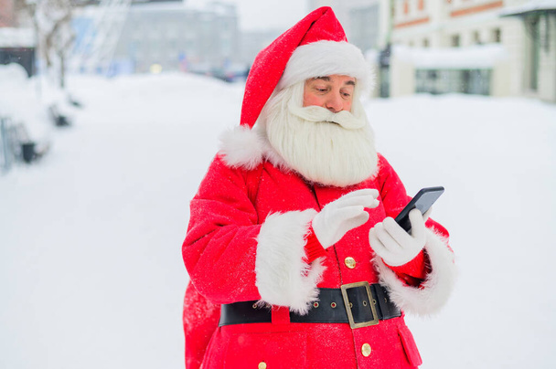Portrait of a funny funky fat modern Santa Claus use his mobile phone. An elderly man in a suit before Christmas communicates on a smartphone. - 写真・画像