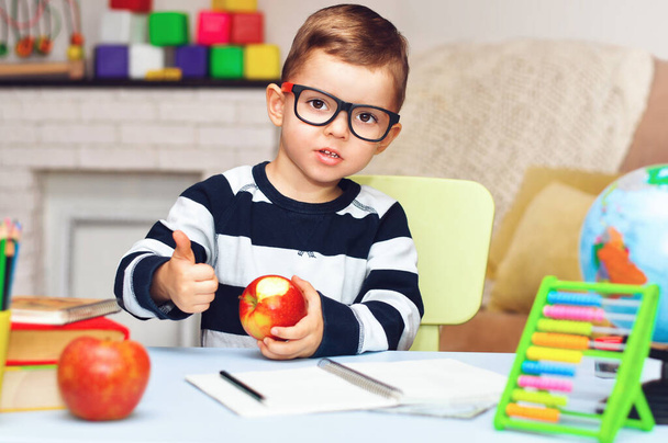 Un niño en edad preescolar es considerado y se sienta en una mesa con un libro, un globo, un cuaderno y lápices y muestra un pulgar hacia arriba
. - Foto, imagen