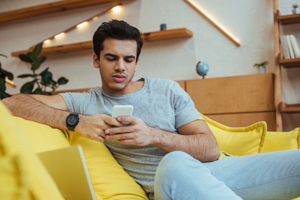 Селективный фокус концентрированного человека чата на смартфоне на диване в гостиной
 - Фото, изображение
