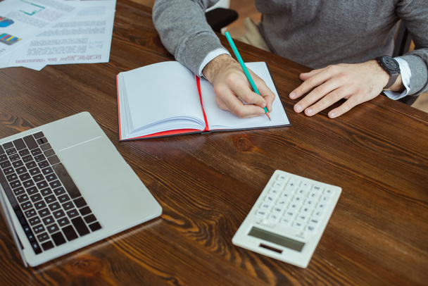 Ausgeschnittene Ansicht eines Geschäftsmannes, der in einem Notizbuch neben Laptop, Taschenrechner und Papieren am Tisch im Büro schreibt - Foto, Bild