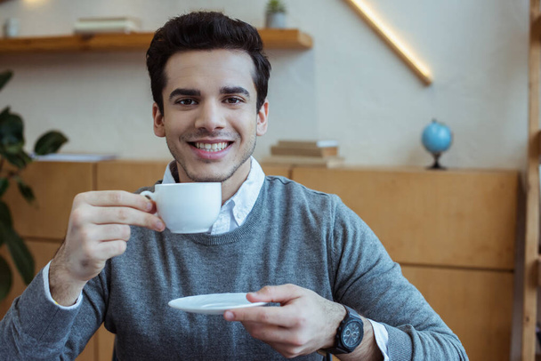 Geschäftsmann hält Untertasse und Tasse Kaffee, lächelt und blickt in die Kamera im Büro - Foto, Bild