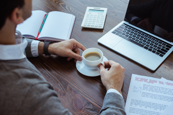 Ausgeschnittene Ansicht eines Geschäftsmannes mit einer Tasse Kaffee in der Nähe von Laptop, Taschenrechner, Papieren und Notizbuch am Tisch  - Foto, Bild